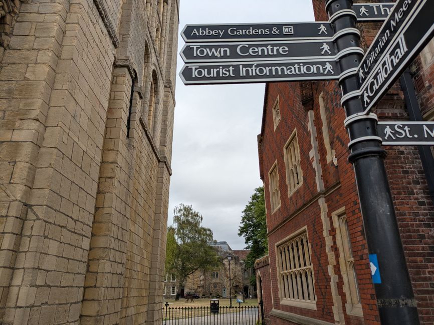 A black street signpost with multiple directional signs mounted on a pole, pointing towards Abbey Gardens & WC, Town Centre, and Tourist Information, situated on a pavement in front of historic buildings. To the left is a stone building with detailed masonry, while on the right, there is a red brick building with decorative window frames. The sky overhead is overcast, casting diffuse lighting on the scene. In the background, a small park or courtyard area is visible through a black metal fence, with trees and a bench. The image context relates to the location near Abbey Road Studios in St Johns Wood, with the signage indicating nearby attractions or amenities. This setting aligns with house removals and relocation services offered by company [COMPANY_NAME], illustrating the urban environment where furniture transport and home moving activities may take place.