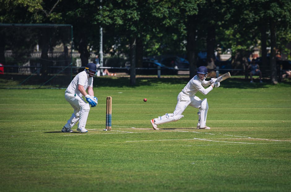 A cricket match taking place on a well-maintained green field, with two players dressed in white cricket uniforms, protective helmets, and gloves. One player is positioned behind the stumps, ready as the wicketkeeper, crouched and prepared to catch or stop the ball. The other player is batting, mid-swing with a wooden cricket bat, aiming to hit a red cricket ball approaching from the right. The grass pitch area is clearly visible, with cricket stumps and bails behind the wicketkeeper. In the background, there are trees and a few spectators seated on benches, indicating an outdoor setting. This scene, captured during daylight hours, illustrates a dynamic moment of a cricket game, relevant to hosting or attending sports events at Lords Cricket Ground, but also connects to the logistics of organising match day access and event support, similar to elements involved in home or venue relocation and access arrangements facilitated by removal services such as those at removalcompaniesstjohnswood.co.uk.